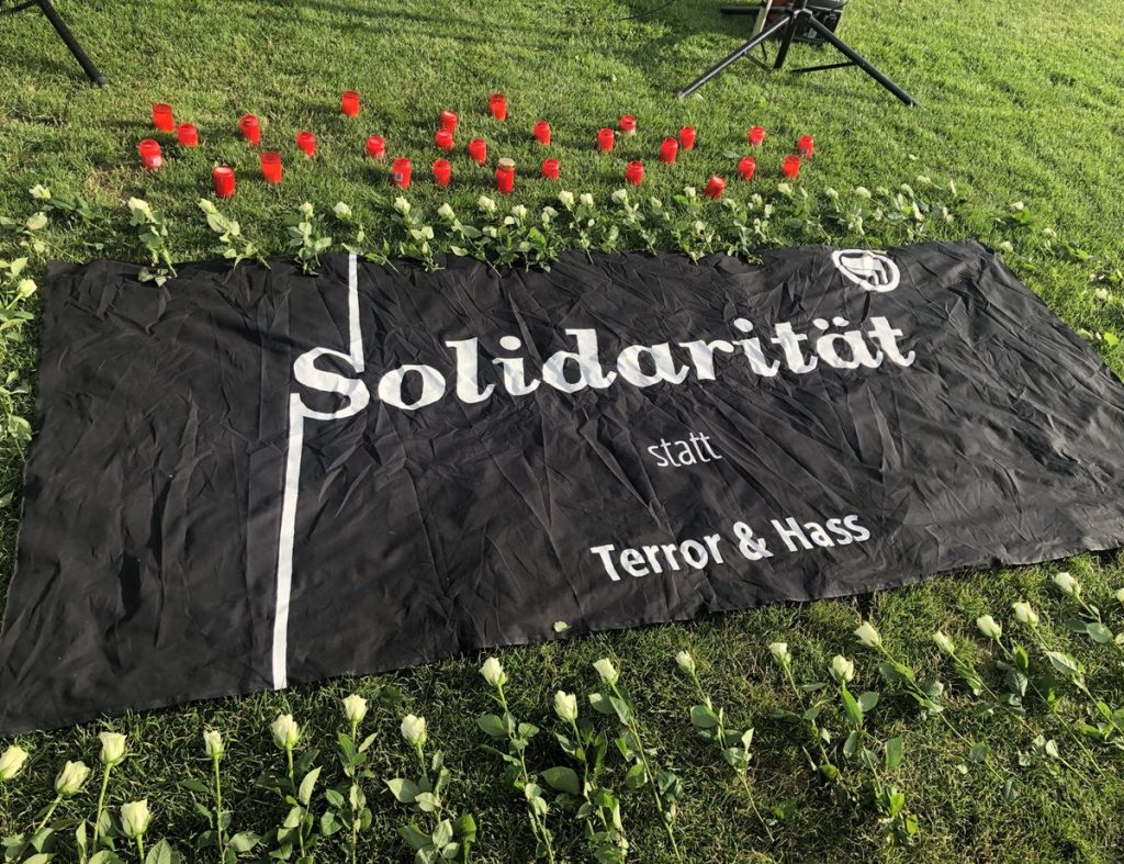 Auf dem in einer Wiese liegenden schwarzen Transpi steht mit weißer Schrift "Solidarität statt Terror & Hass". Im rechten oberen Eck des Transpis ist eine weiße Antifa Flagge gezeichnet. Um das Transpi liegen Blumen, hinter dem Transparent stehen Grabkerzen